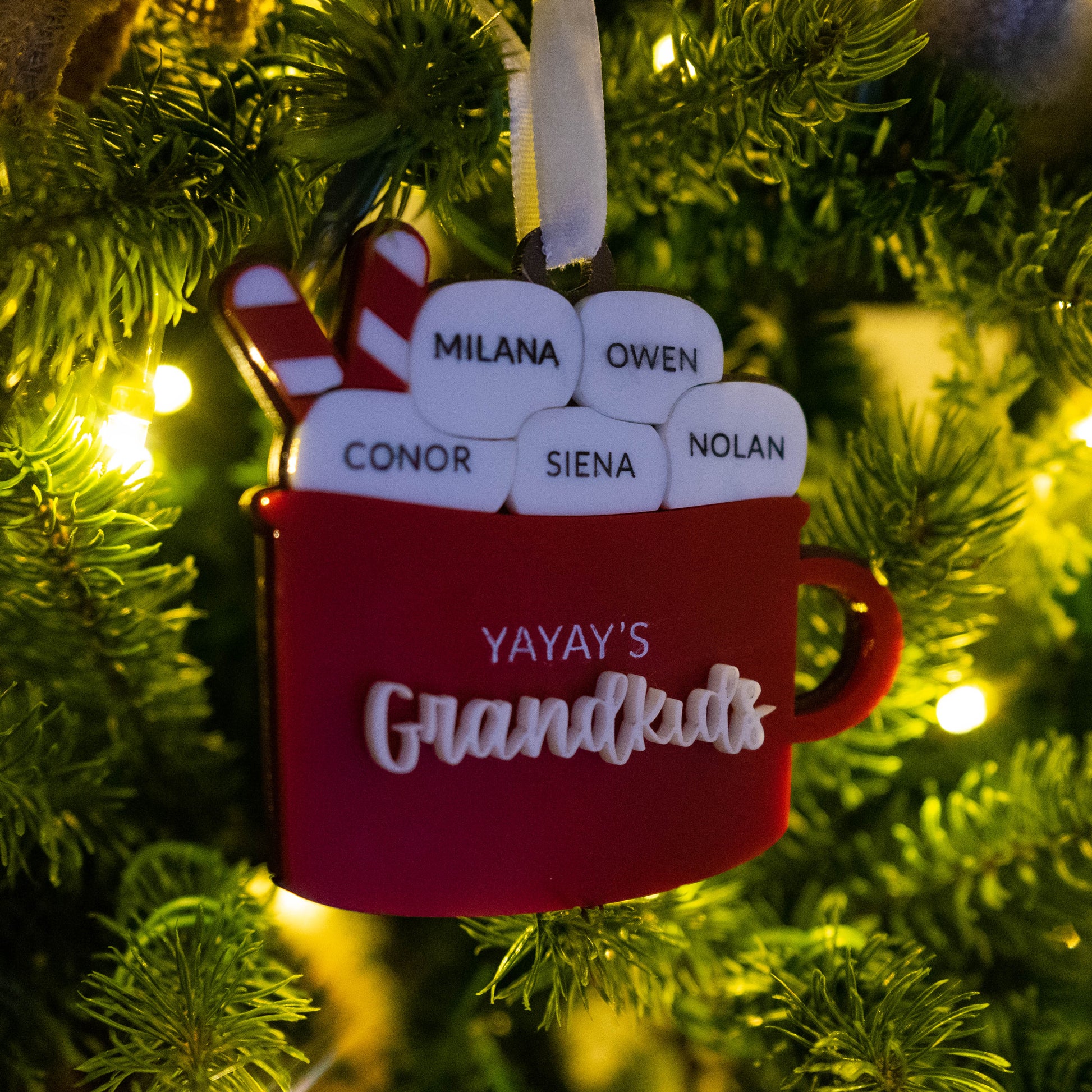 Personalized hot cocoa ornament on a Christmas tree with marshmallows showing grandkids’ names and a Bible verse engraved on the back, Christian grandparent keepsake.