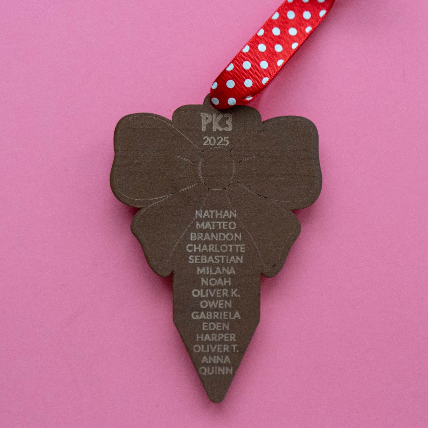 Engraved wood ornament back showing student names under “PK3 2025,” red polka-dot ribbon, pink background.