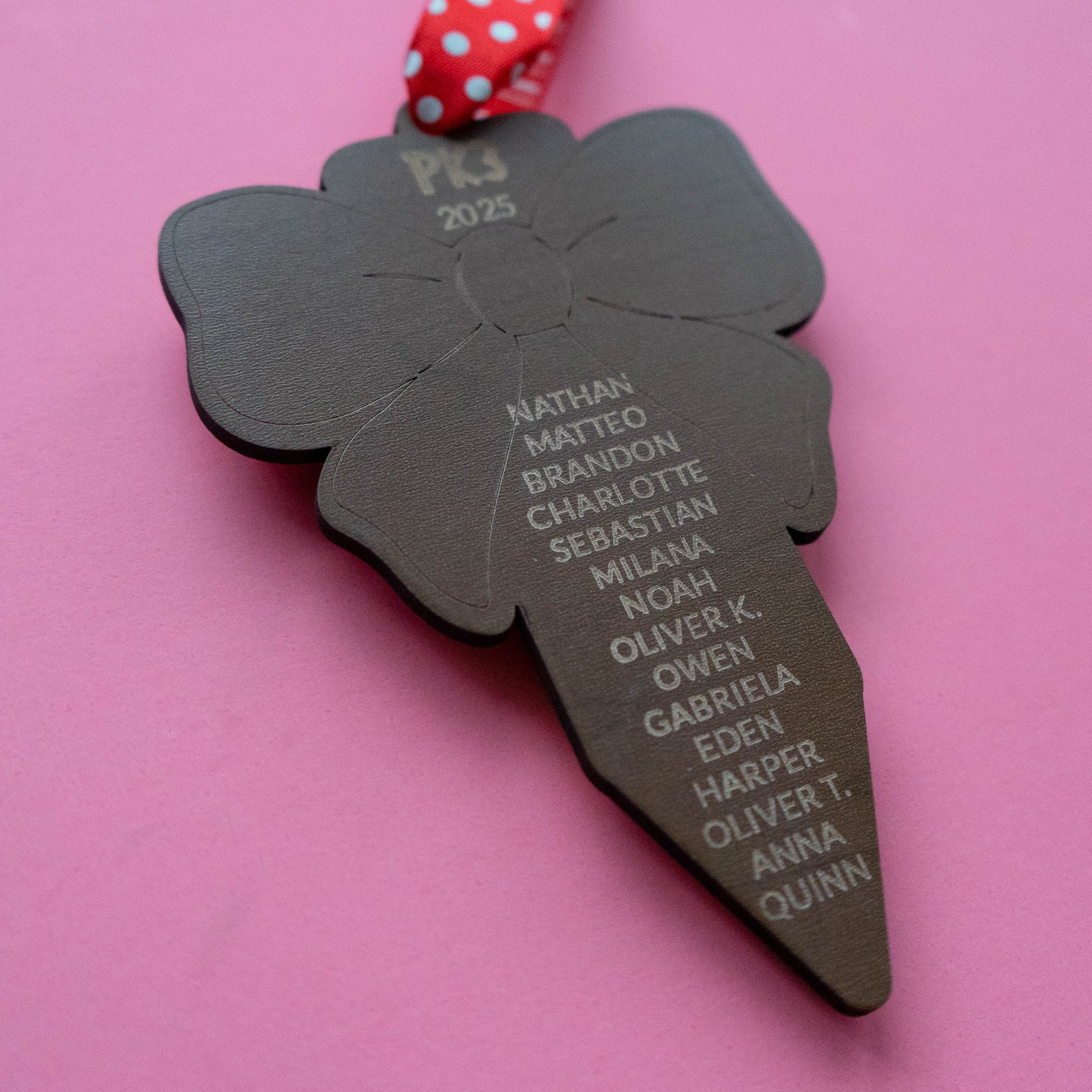 Engraved wood ornament back showing student names under “PK3 2025,” red polka-dot ribbon, pink background.