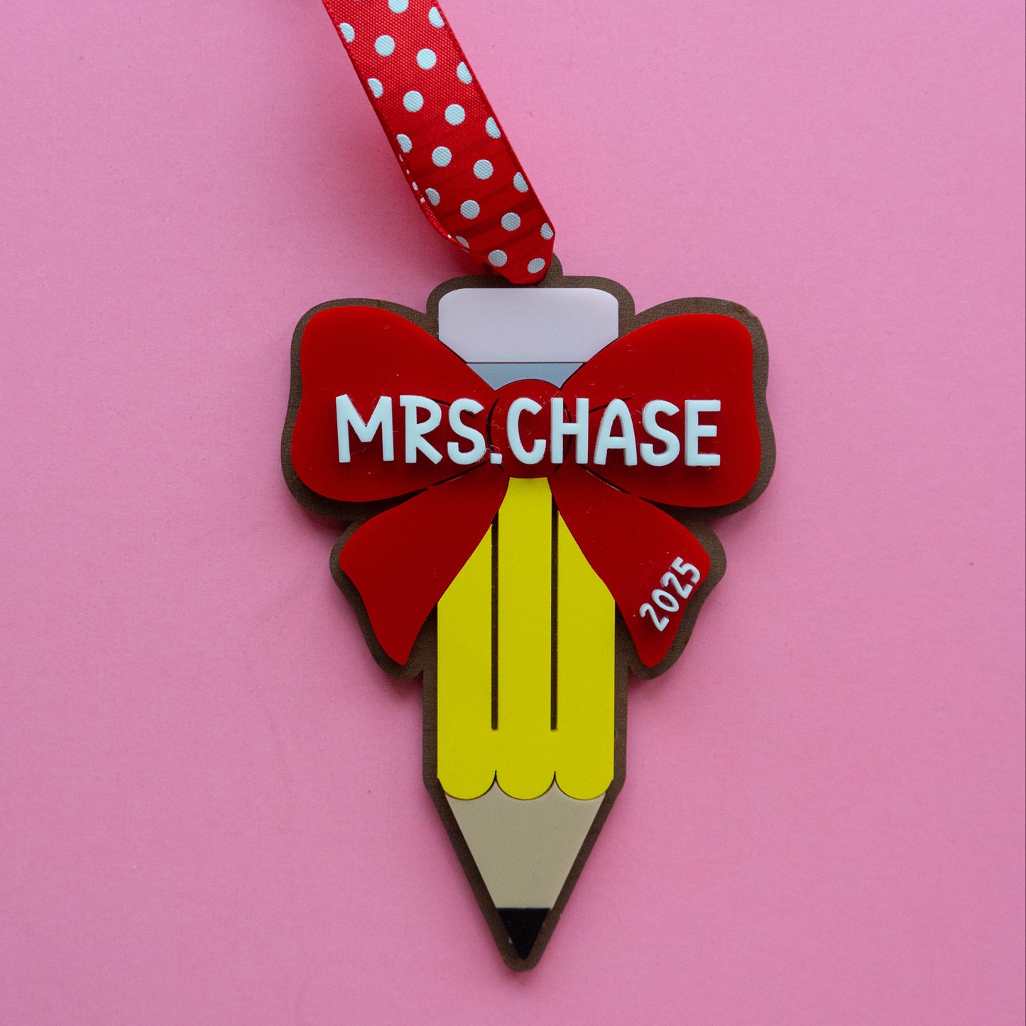 Personalized teacher pencil ornament with red bow, teacher name “Mrs. Chase,” red polka-dot ribbon, pink background.