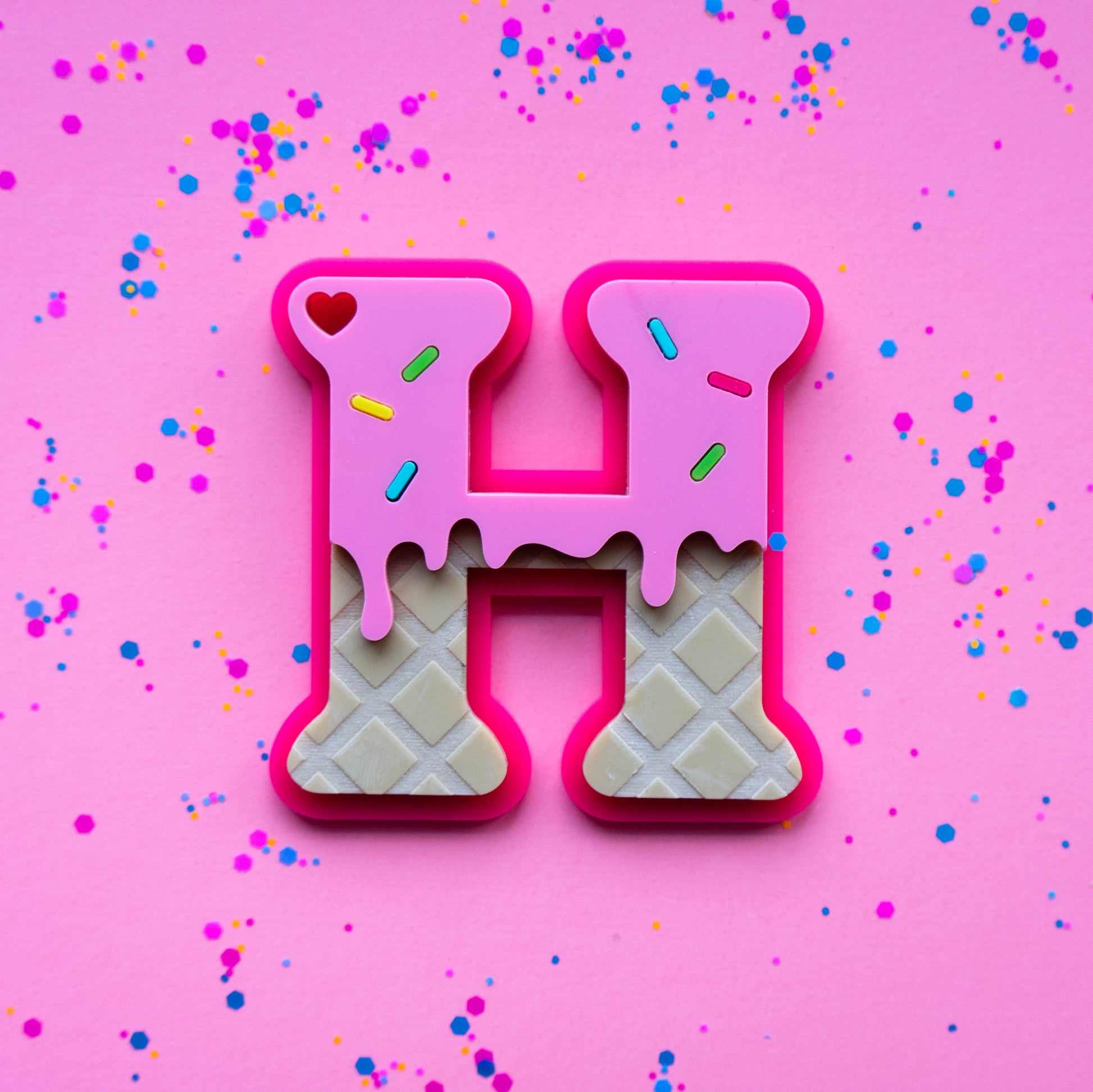 Letter 'H' acrylic bag tag made of waffle with pink icing on a pink background with confetti.