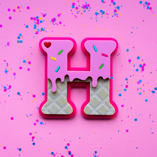 Letter 'H' acrylic bag tag made of waffle with pink icing on a pink background with confetti.