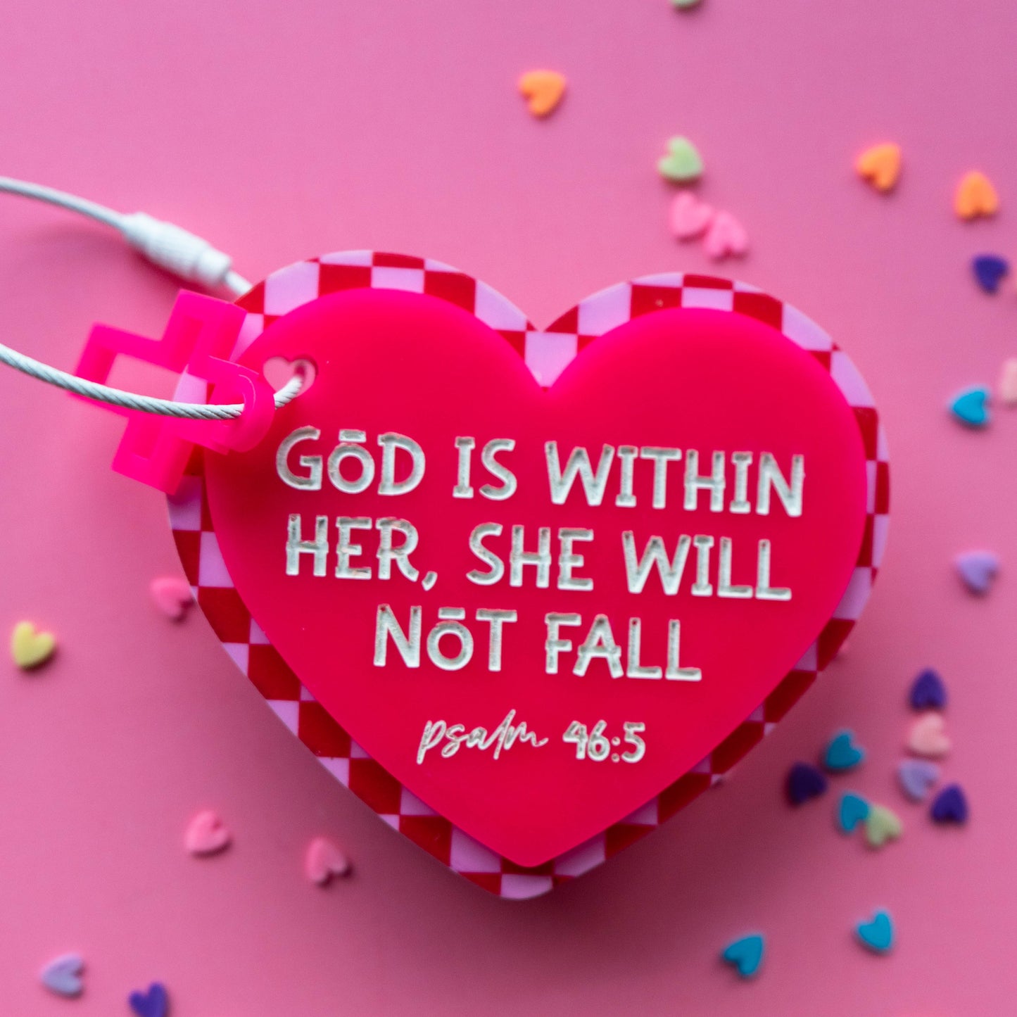 Heart-shaped acrylic bag tag with 'God is within her, she will not fall' text on a pink background with heart-shaped confetti.
